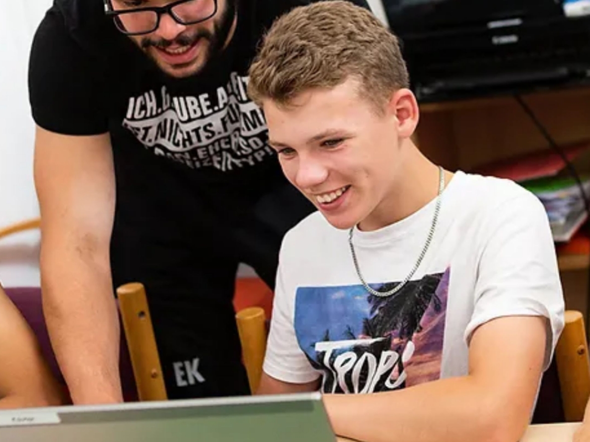 Ein Betreuern schaut seinem Schützling, der am Laptop sitzt, über die Schulter