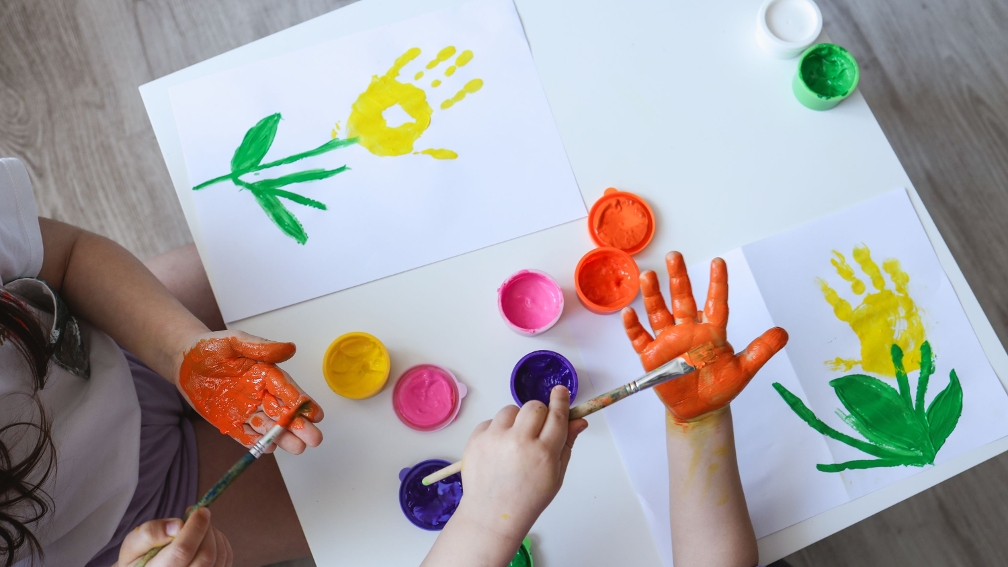 Kinderhände, die auf ein Blatt Papier ihre Handabdrücke mit bunter Farbe setzen